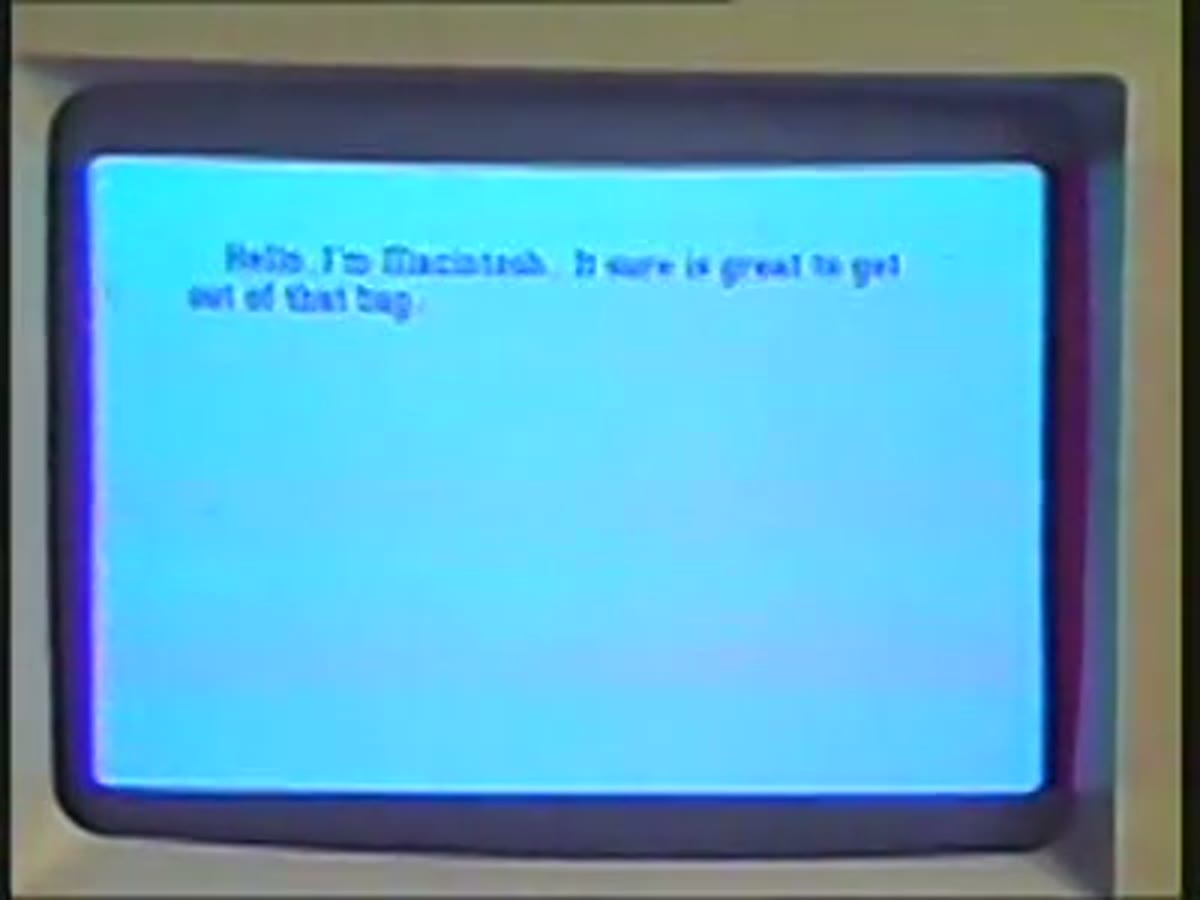 A blue screen reading “Hello, I am Macintosh. It sure is great to get out of that bag.”